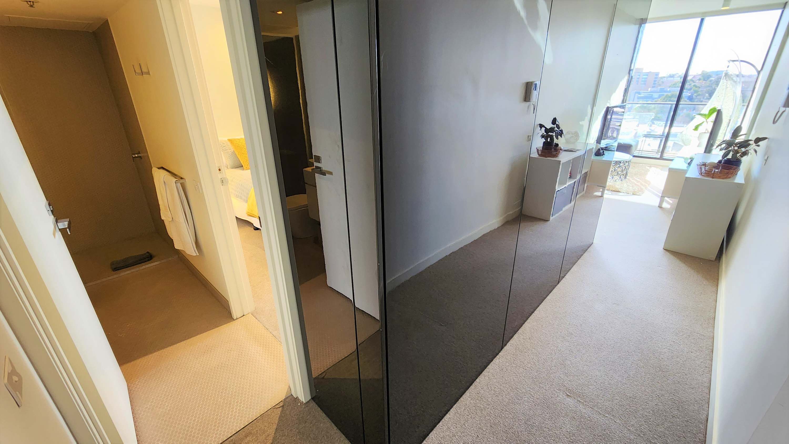 A modern hotel corridor with a glass partition, a bed, a small desk, and a view of the city through large windows.