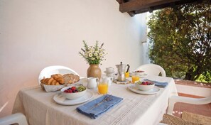 A breakfast table set outdoors with croissants, a fruit bowl, a vase with flowers, a coffee maker, and a pitcher of orange juice.