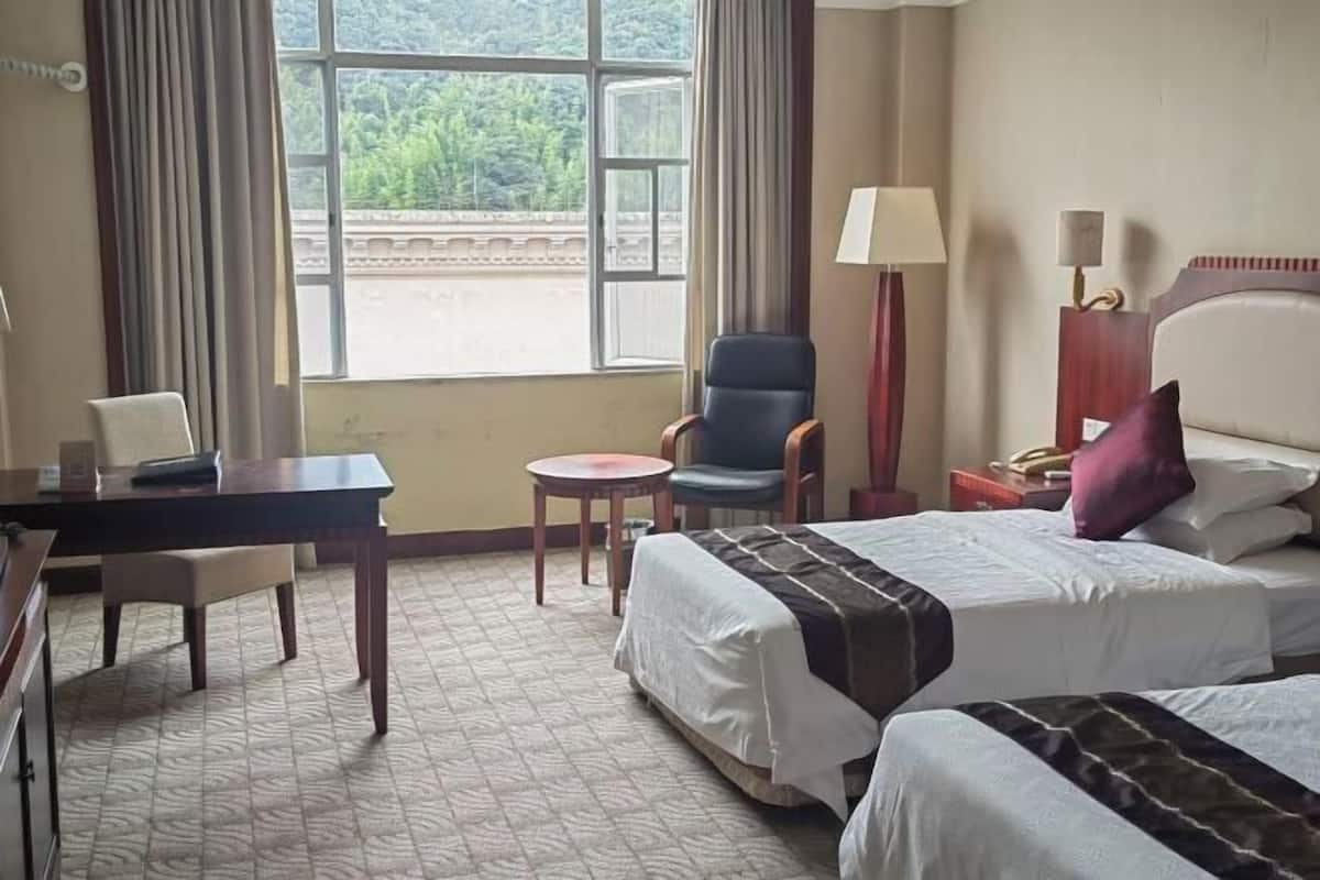 A hotel room with two beds, a desk, a chair, a lamp, and a view of greenery outside.