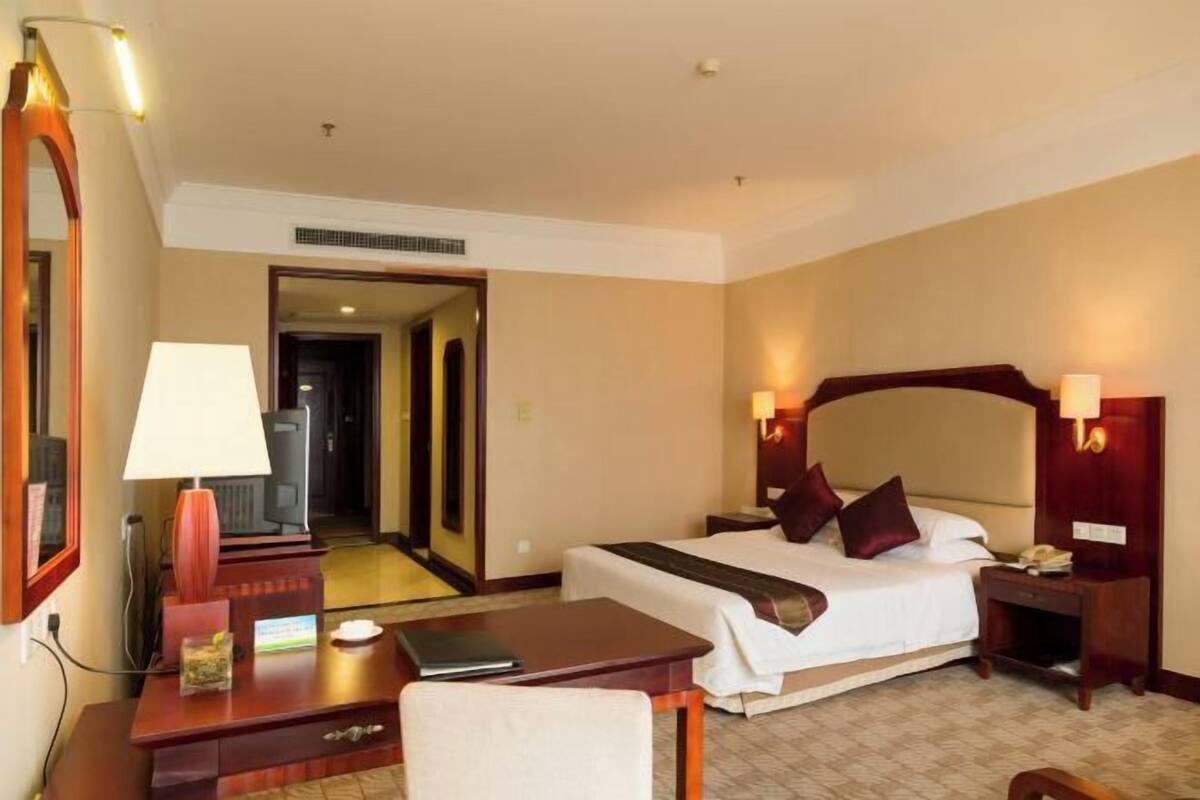 A hotel room with a bed, bedside tables, a desk, a chair, a lamp, and a mirror.