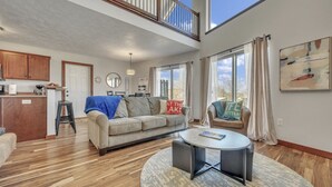 A spacious living area with a high ceiling, a kitchen in the background, and a large window with curtains.