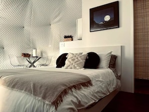 Un dormitorio moderno con techo acolchado, una cama con varias almohadas, una mesita de noche con lámpara y un cuadro enmarcado en la pared.