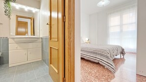 A bathroom with a vanity, mirror, and a door leading to a bedroom with a bed and a window.