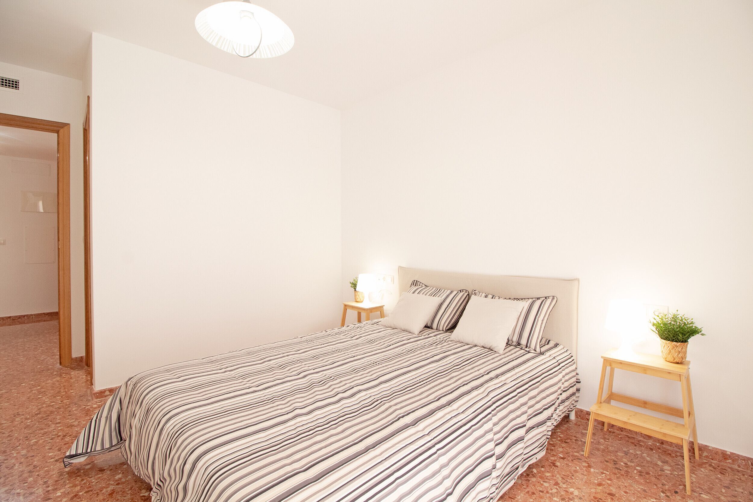 Een slaapkamer met een gestreept beddegoed, een nachtkastje met een lamp en een kleine plant op een bijzettafeltje.