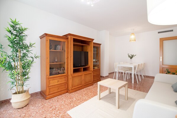 A living room with a wooden cabinet, a TV, a dining table with chairs, a white sofa, and a potted plant.