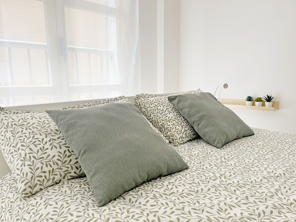 A bed with patterned bedding and pillows, a window with sheer curtains, and a small shelf with potted plants.