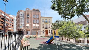 A playground with a slide and benches, surrounded by a fence, with buildings in the background.