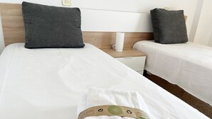 Ein Hotelzimmer mit zwei Einzelbetten, jeweils mit einem grauen Kopfkissen und weißer Bettwäsche. Auf dem Bett im Vordergrund liegt ein weißes Handtuch mit einem grün-braunen Etikett.