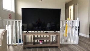 Una sala de estar con televisor de pantalla plana sobre un mueble de madera, un candelabro y una barandilla blanca con detalles amarillos.