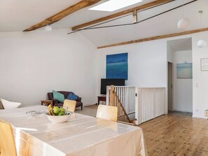 Ein großzügiger Wohnbereich mit hoher Decke, Holzbalken, einem Esstisch mit Obstschale, einer Couch mit Kissen und einem Gemälde an der Wand.
