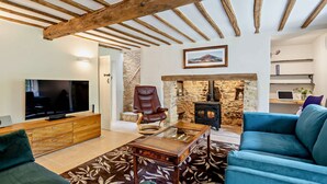 A living room with exposed wooden beams, a stone fireplace, a television, a leather armchair, and a blue sofa.