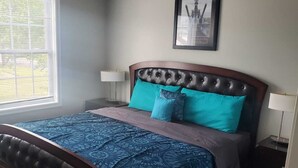 A bedroom with a wooden bed, a window with blinds, a framed picture on the wall, and a ceiling light. - Welcome to our newly built house  (Glen Burnie)