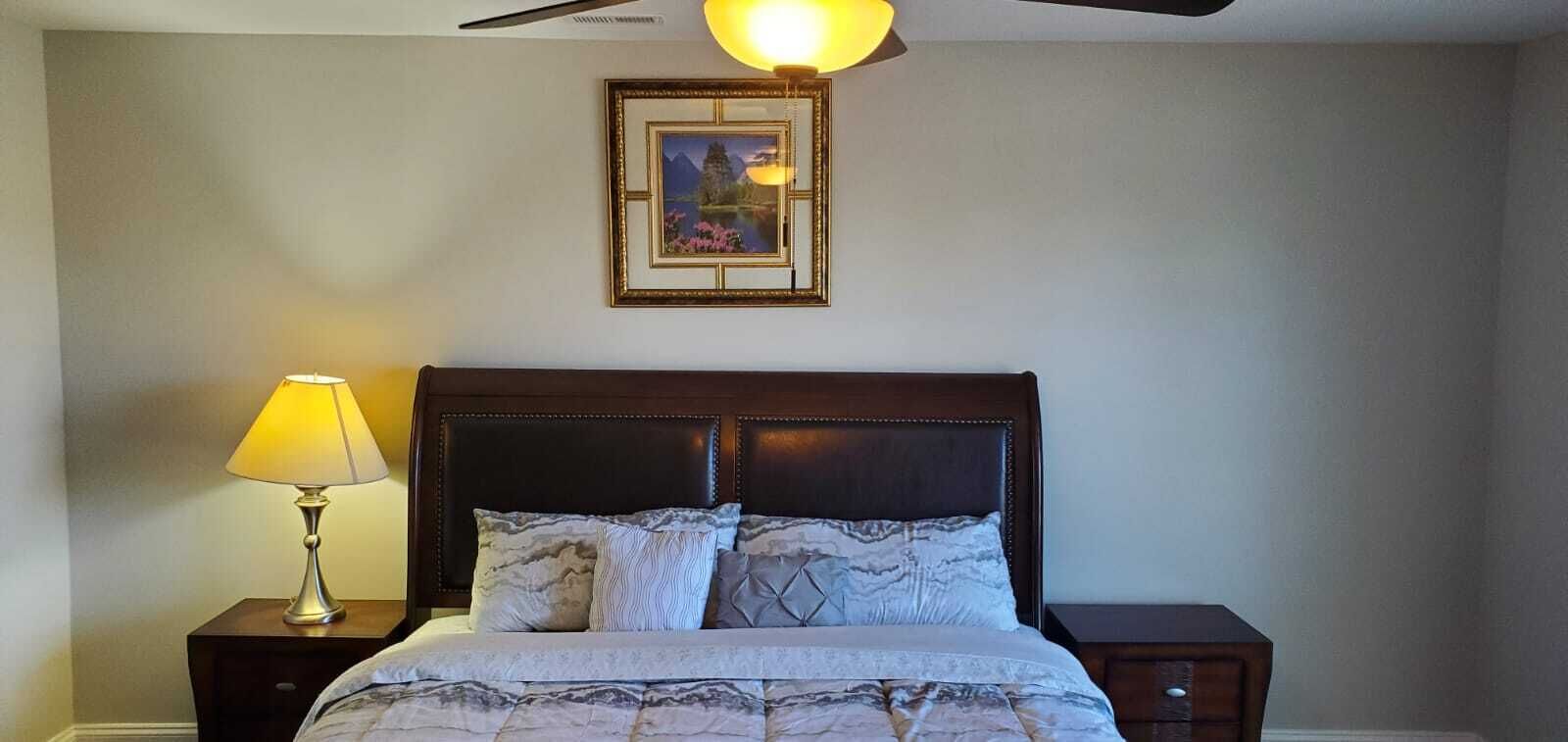 A bedroom with a bed, a nightstand, a lamp, a framed picture on the wall, and a ceiling fan.
