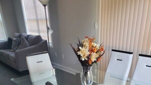 A modern dining area with a glass-top table and white chairs, a vase of flowers, and a grey sofa in the background. - Welcome to our newly built house  (Glen Burnie)