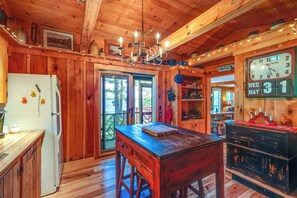 A wooden cabin interior with a kitchen area, wooden walls, a chandelier, a clock, and a dining table. - Waterfront Maine Vacation Rental w/ Private Dock (Bradley)