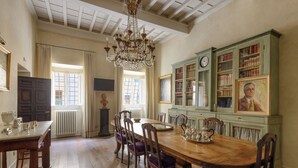 A dining room with a wooden table, chairs, a chandelier, and a green bookcase with books and a clock.