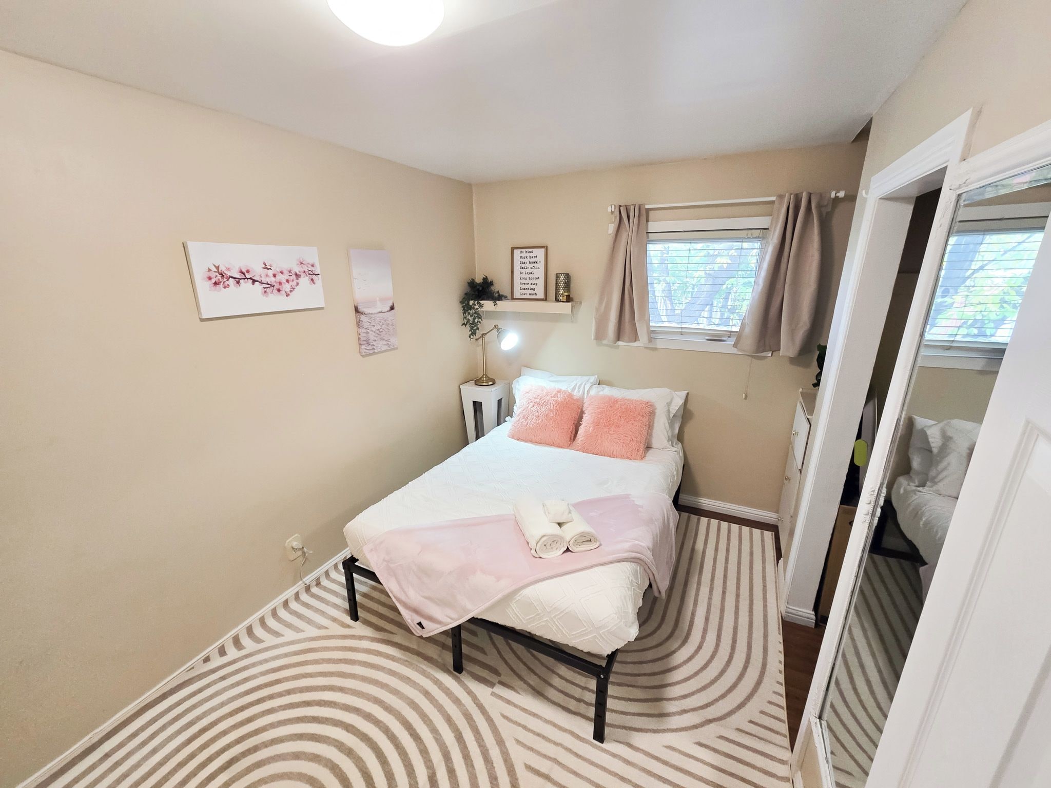 A bedroom with a bed, a bedside table, a lamp, a window with curtains, and a wall with framed pictures.
