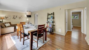 A dining area with a wooden table set for four, a bookshelf, and a sofa in a well-lit room with wooden floors. - Family House with Httb Playgr BBQ Firept (Calgary)