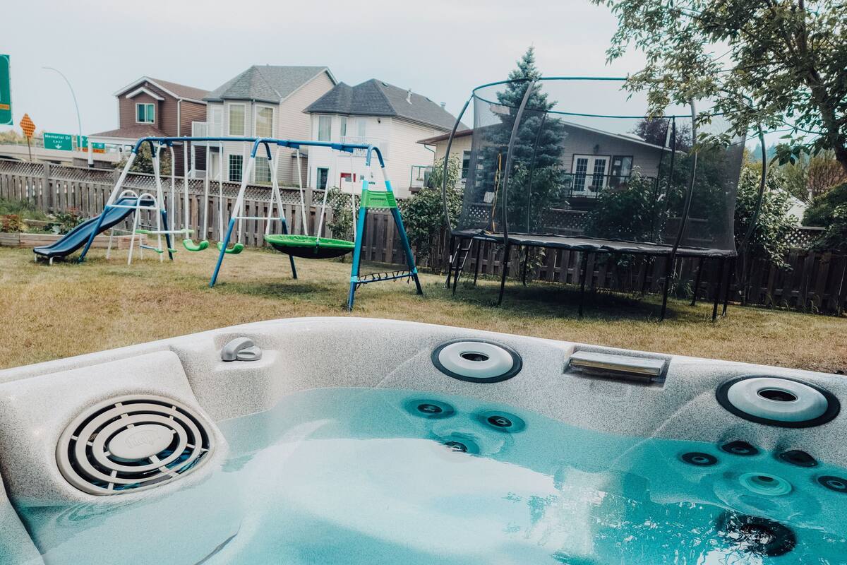 A hot tub with a view of a backyard with a swing set and a trampoline.