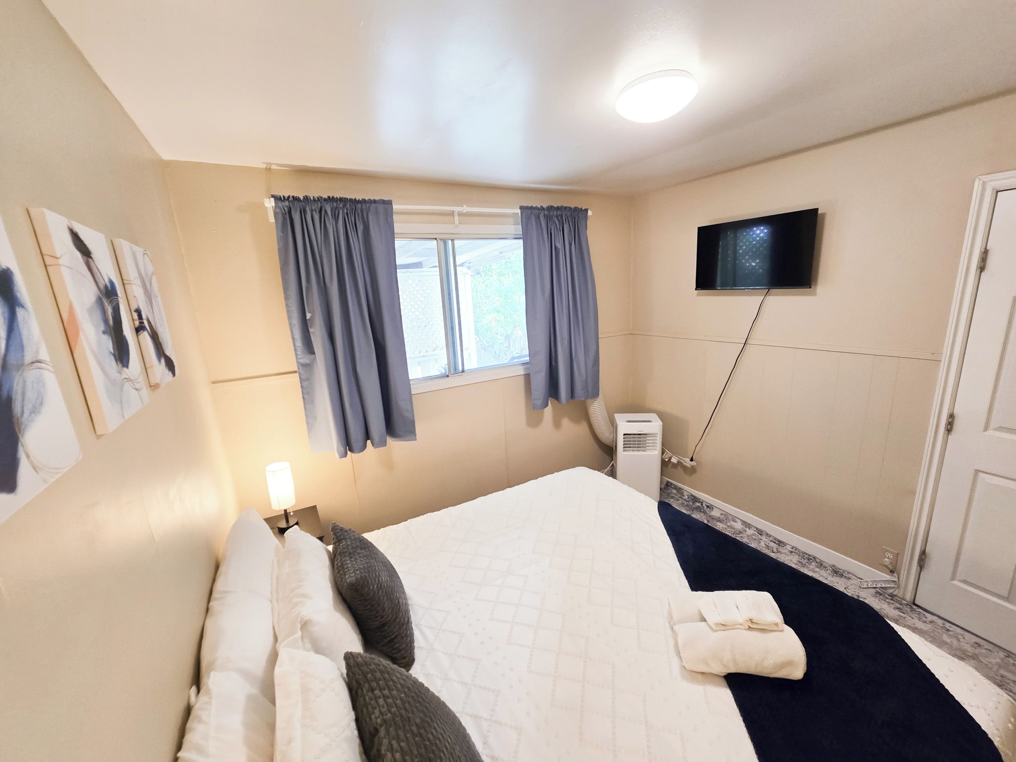 Een hotelkamer met een bed, een flatscreen televisie aan de muur, een raam met gordijnen, een bedlamp en een airconditioner.