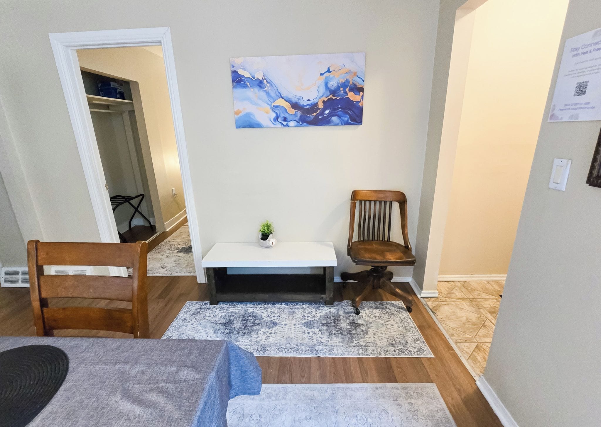 Een kamer met een houten stoel, een kleine tafel met een plant, een blauw-wit gestreept tapijt en een schilderij aan de muur.