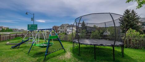 A playground with a swing set and a trampoline, surrounded by a wooden fence and houses in the background.