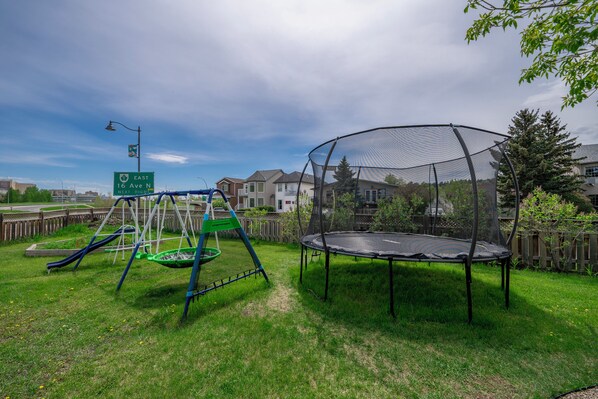 A playground with a swing set and a trampoline, surrounded by a wooden fence and houses in the background. - Family House with Httb Playgr BBQ Firept (Calgary)
