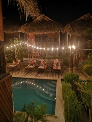 Un espace aménagé au bord de la piscine, avec des chaises longues en bois et des structures à toits de chaume, le tout entouré de verdure.