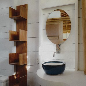 Une salle de bain avec une étagère en bois, un miroir rond et un lavabo bleu.