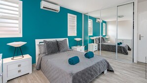 A modern bedroom with teal walls, a large bed with grey bedding, two bedside tables with lamps, and a large mirror.