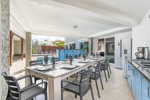 Ein Essbereich mit einem langen Tisch, gedeckt für sechs Personen, eine Küche mit modernen Geräten und Blick auf den Pool sowie angrenzende Gebäude.