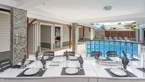 A dining area with a white table set for four, a blue pool in the background, and a stone wall on the left.