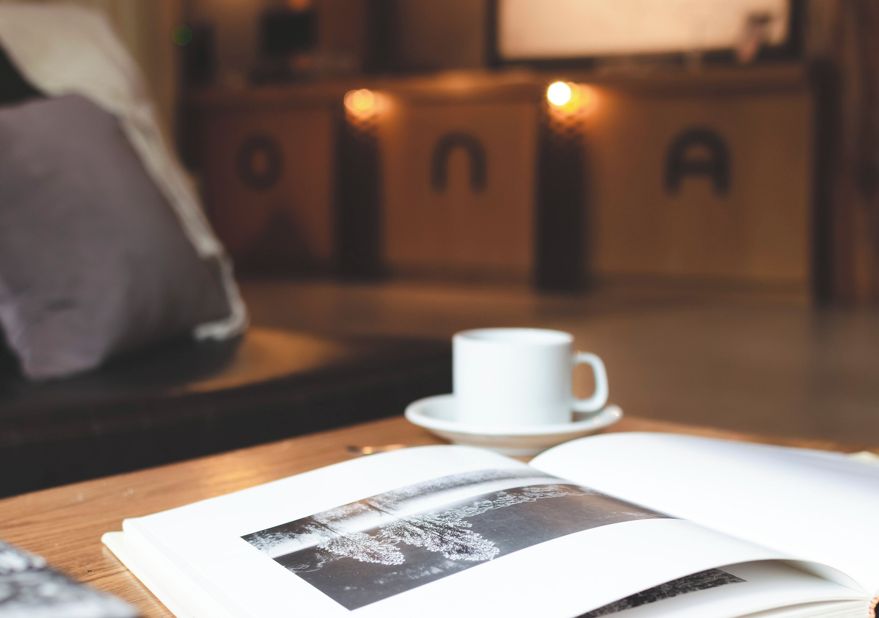 Un rincón acogedor para leer con una taza blanca sobre un platillo, un libro y un cojín sobre una mesa de madera.