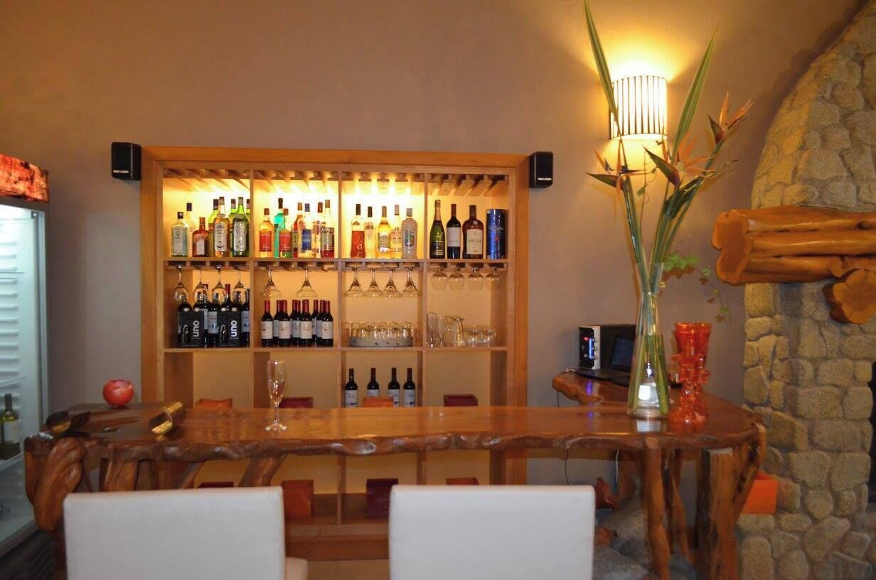Una barra de madera con estantes llenos de botellas de licores y vasos, una pared de piedra y una planta alta.