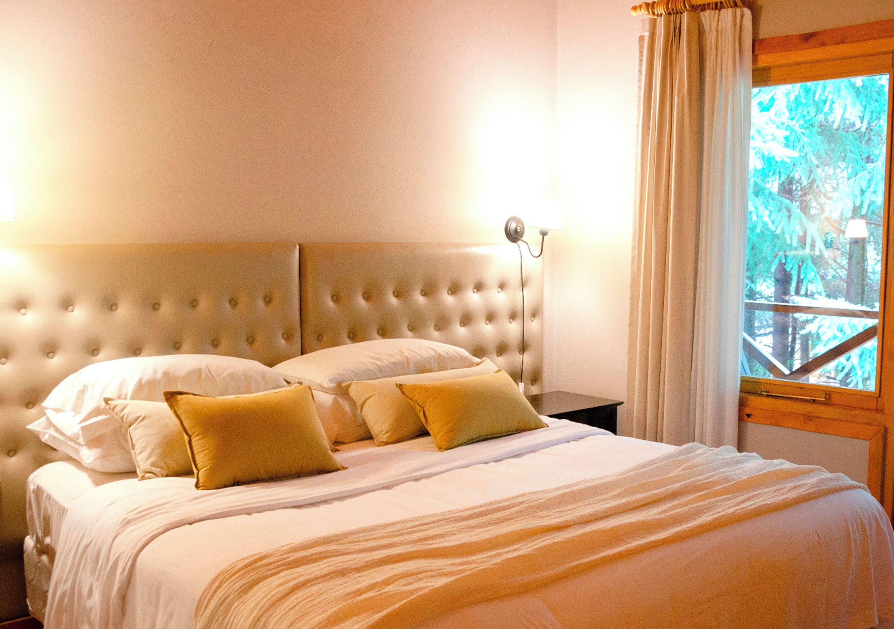 Een slaapkamer met een knopen hoofdbord, een bed met wit en mosterdkleurig beddengoed, en een raam met uitzicht op bomen.