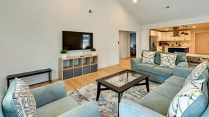 Una sala de estar moderna con un sofá, una mesa de centro y un televisor. Se ve un área de cocina al fondo.