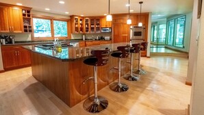 Cozinha moderna com armários de madeira, uma ilha grande com banquetas e vista para o exterior através de amplas janelas.