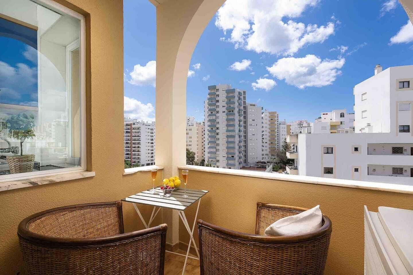 Ein Balkon mit Korbstühlen, einem kleinen Tisch mit Getränken und Blick auf hohe Gebäude.