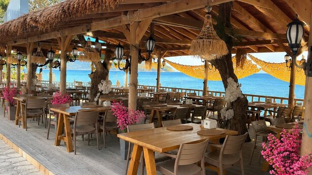 A wooden outdoor dining area with tables and chairs, a thatched roof, and a view of the sea. - Avlu Butik Otel Palamutbükü (Datça)