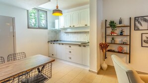 A modern kitchen with white cabinets, a dining table with chairs, and a hanging pendant light. - Avlu Butik Otel Palamutbükü (Datça)