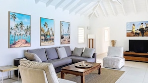 A modern living room with a sectional sofa, a coffee table, and a television. There are paintings on the walls and a ceiling fan. - Villa Eskarcel, très spacieuse, vue mer sur Orient Bay, 4 chambres (Mont Vernon)
