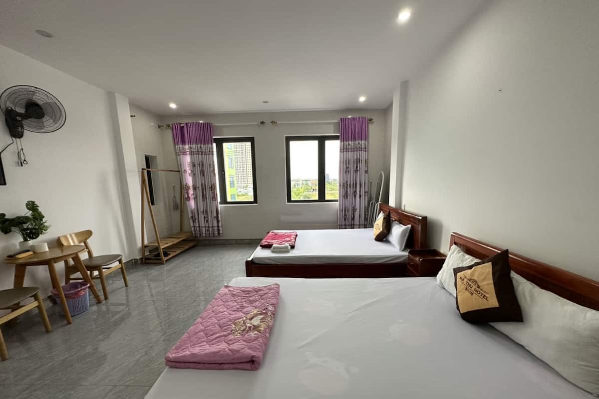 A hotel room with two beds, a desk with chairs, a standing fan, and a window with curtains.