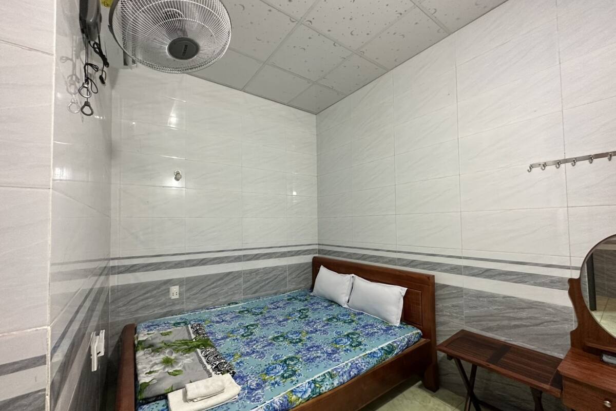 A small, clean room with a bed, a wooden nightstand, a mirror, and a ceiling fan.