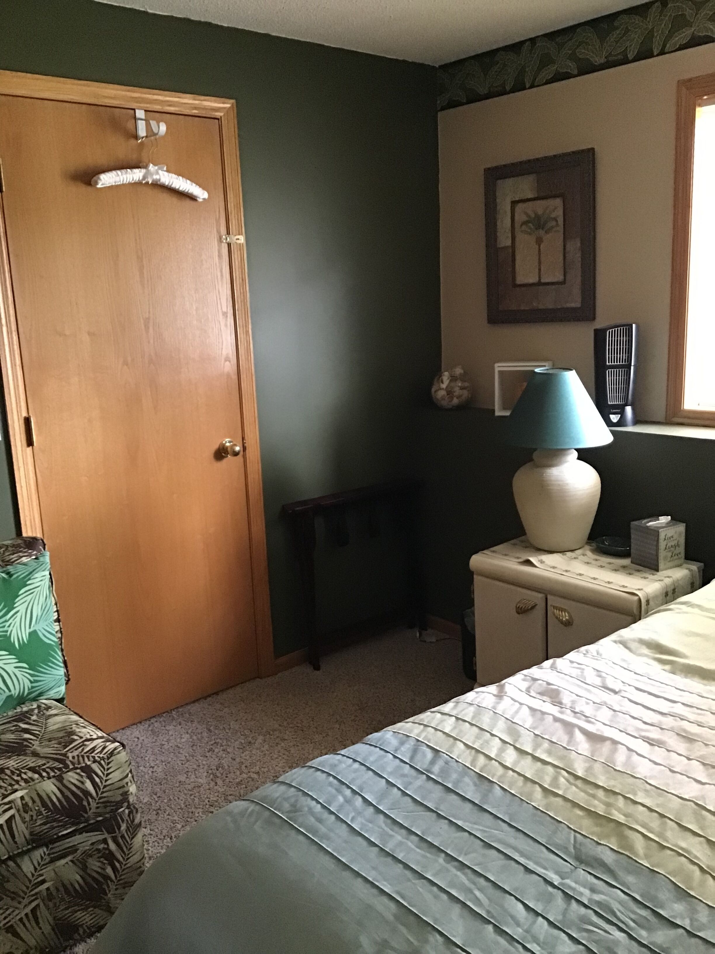 A bedroom with a wooden door, a nightstand with a lamp, a framed picture on the wall, and a bed with a patterned pillow.