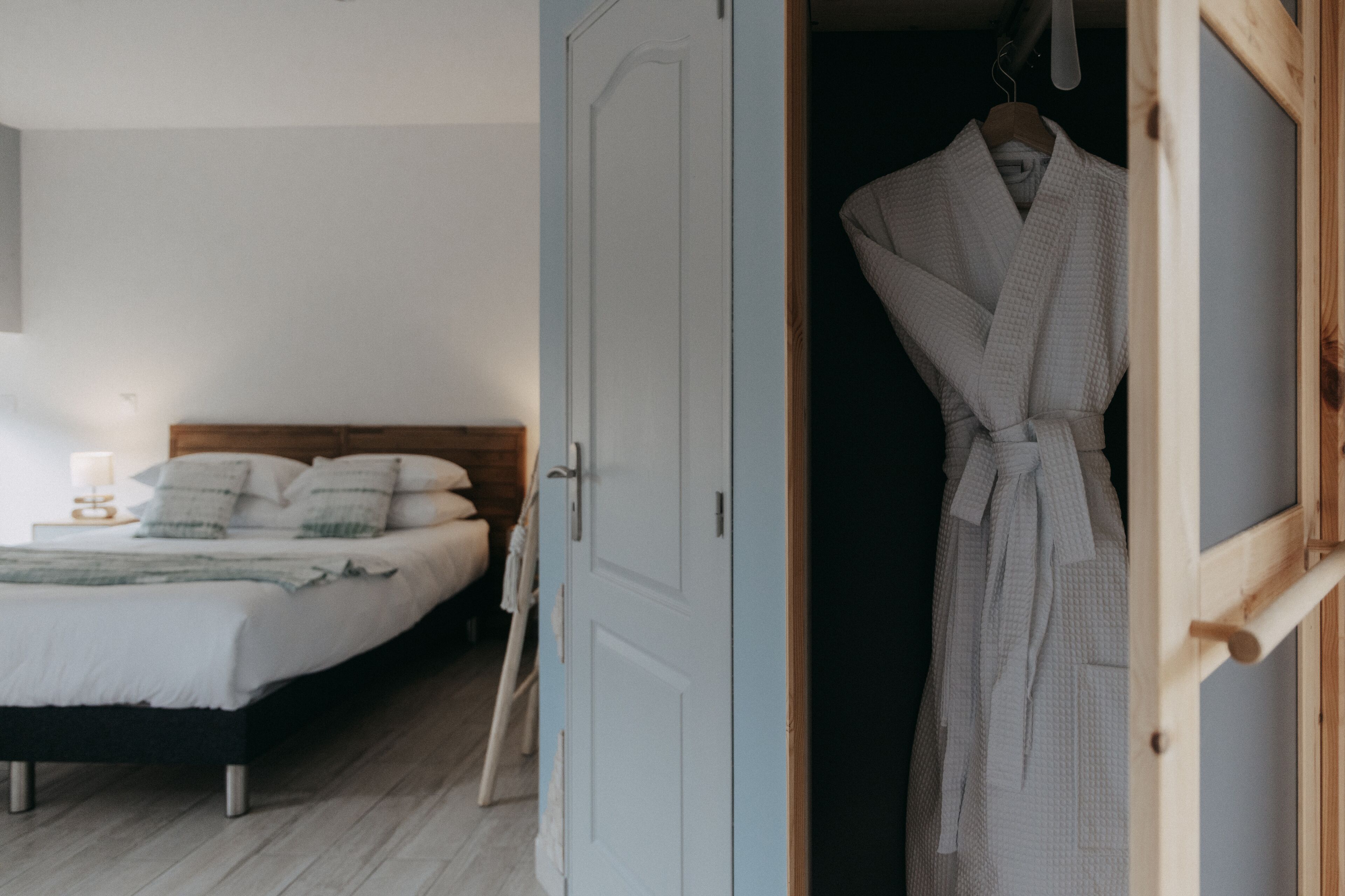 Un dormitorio con una cama, una mesita de noche con lámpara, y una puerta de madera que da a un baño con una bata colgada.