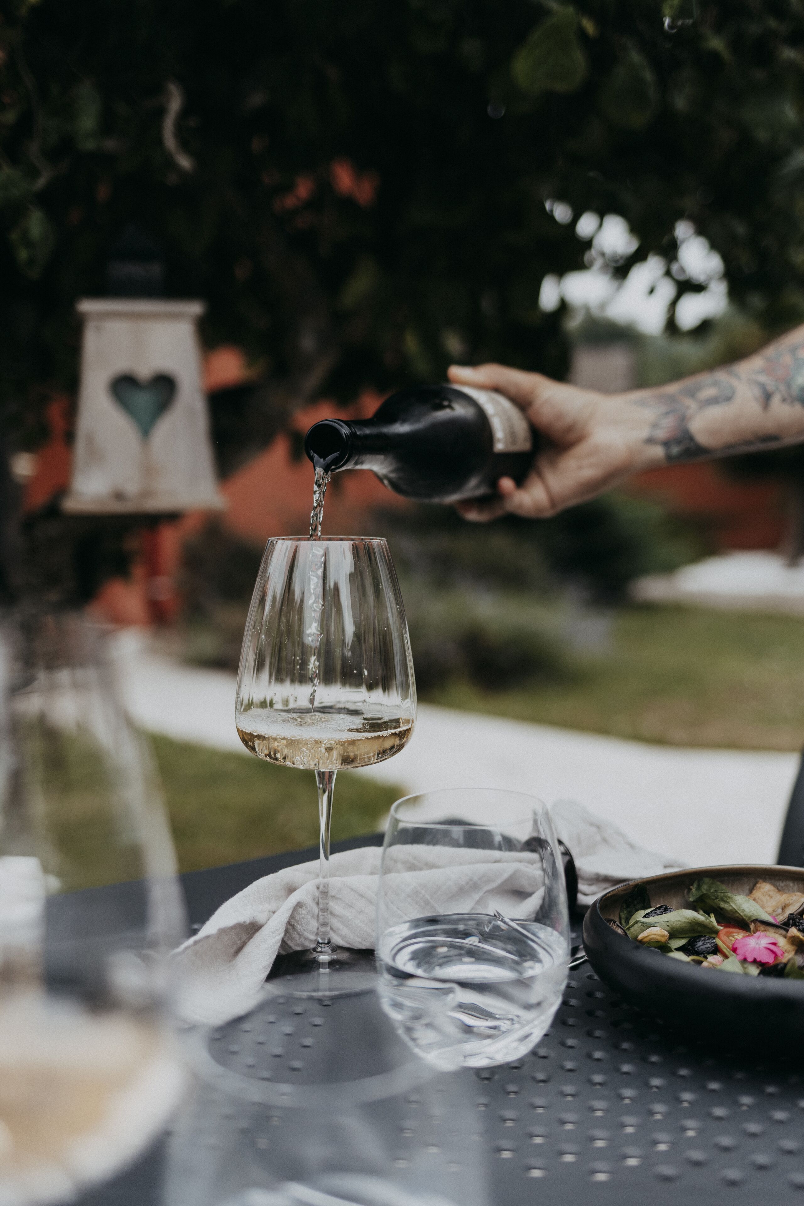 Una copa de vino blanco siendo servida desde una botella, una copa de agua y un bol de ensalada sobre una mesa.