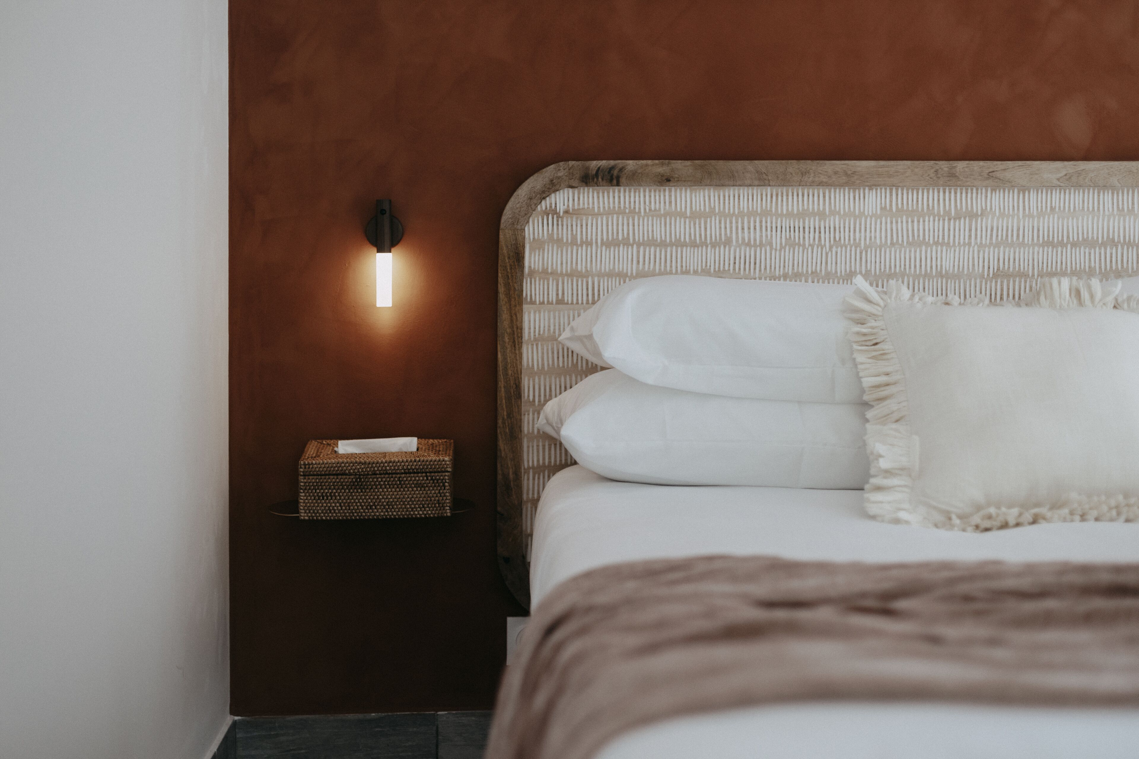 A neatly made bed with white linens and a brown throw blanket, a bedside lamp, and a small wicker basket on a nightstand.