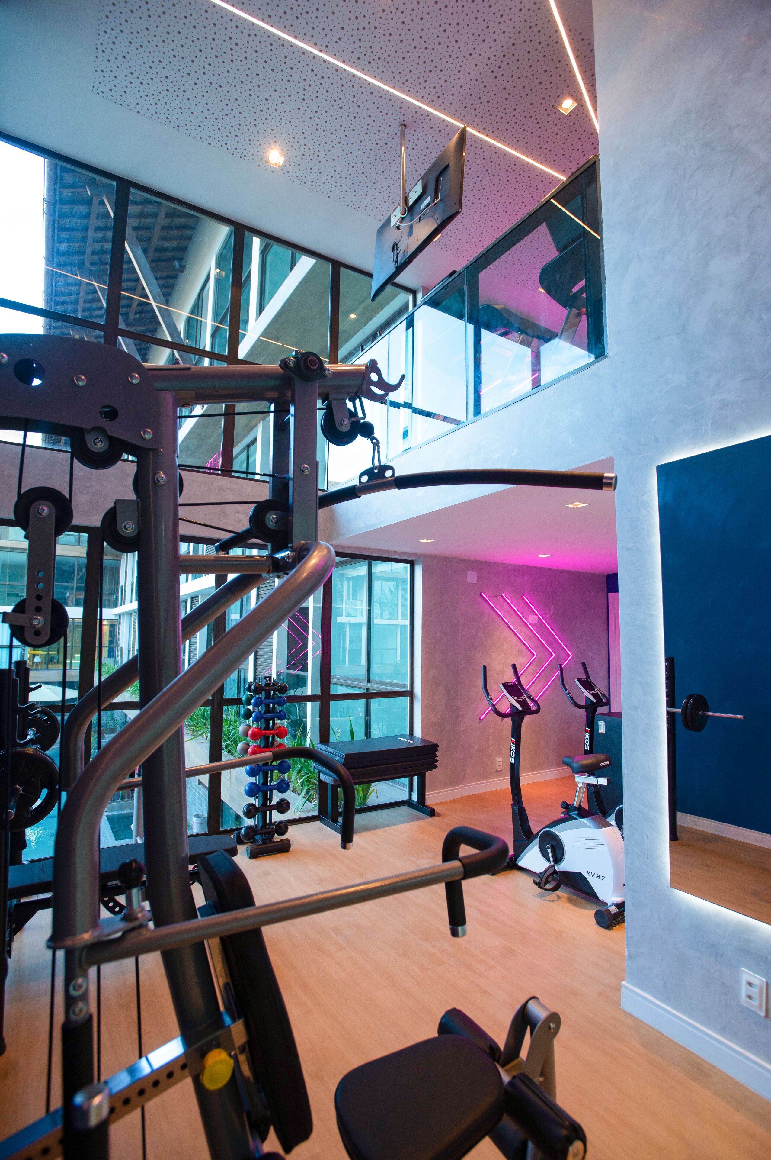 Ein mehrgeschossiges Fitnessstudio mit verschiedenen Trainingsgeräten, großen Fenstern und einer erhöhten Zwischenebene.