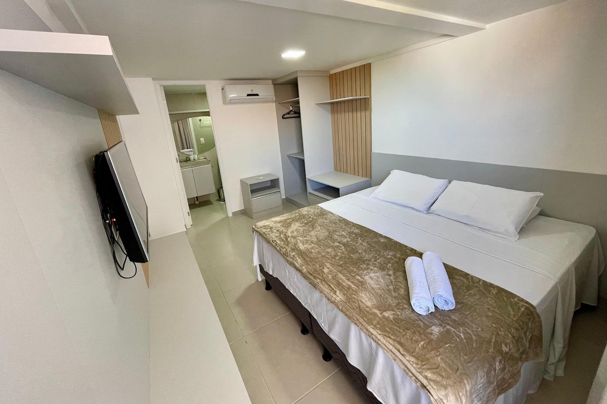 A hotel room with a bed, a TV, a desk, and a bathroom visible through an open door.