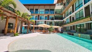 Et moderne hotel med swimmingpool, udendørs siddepladser og en høj bygning med store vinduer.